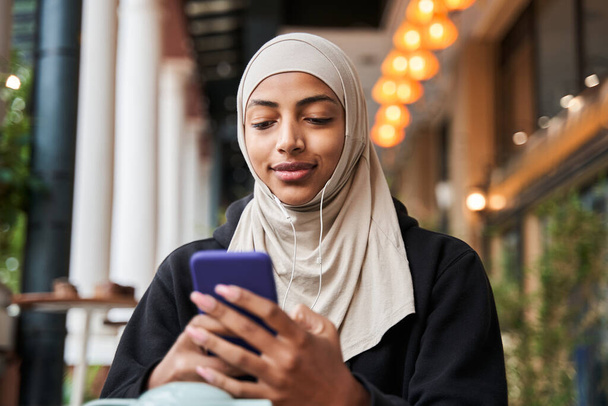 Lady in hijab holding mobile phone and looking at the screen with smile while listening music - Фото, зображення