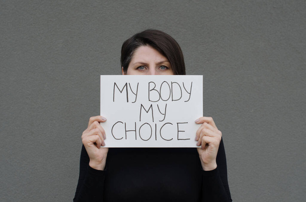 Mujer mostrando pancarta de protesta con el mensaje de mi cuerpo mi elección. Mujer adulta cubre su póster de la cara. Derecho al aborto. Detener la discriminación. - Foto, imagen