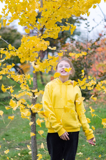 Un adolescente con un suéter amarillo sobre un fondo de hojas amarillas. La chica arroja las hojas. La muchacha se alegra en otoño. Marco vertical. - Foto, Imagen