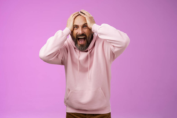 Outraged furious mad adult man bearded yelling shocked despair hatefully looking camera insane holding hands head frowning shouting losing bet disappointed very upset standing purple background - Фото, зображення