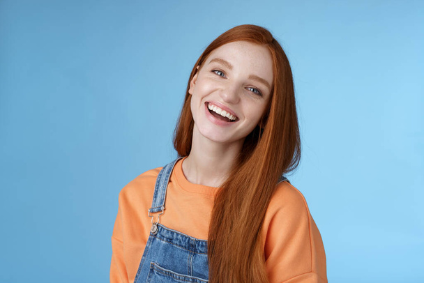 Positive outgoing lively redhead girl laughing joyfully having fun talking friendly friends tilting head chuckling joking funny life moments standing positive lucky blue background orange t-shirt - Фото, зображення