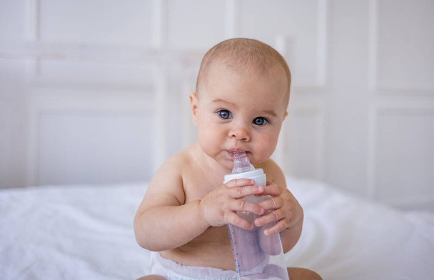 retrato de una niña sentada y bebiendo un biberón de agua en una cama blanca en una habitación. Cuidado de niños - Foto, Imagen