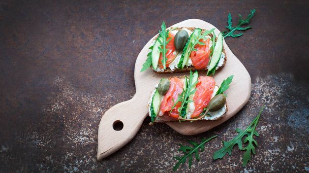 Rye bread sandwiches with trout, cream cheese, capers, cucumbers, arugula on a wooden board on dark brown background. Top view - Photo, image
