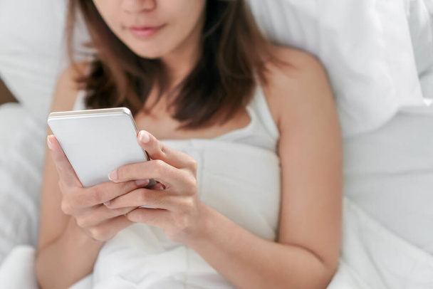 Young asian woman playing with her smathphone on white pillow on bed in bedroom on morning time after wake up, Selective focus. - Foto, Bild