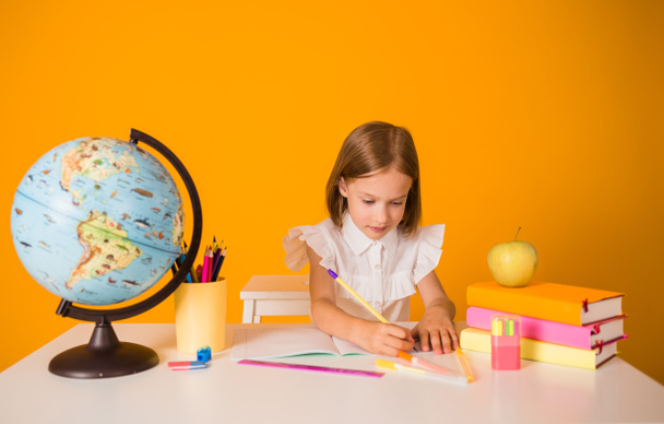 colegiala en una blusa blanca se sienta en una mesa con útiles escolares y un globo y escribe en un cuaderno sobre un fondo amarillo con una copia del espacio - Foto, Imagen