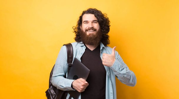 Sonriente joven estudiante en ropa casual con mochila aislada en el estudio de fondo amarillo. Sostenga el ordenador portátil, mostrando el pulgar hacia arriba. - Foto, Imagen