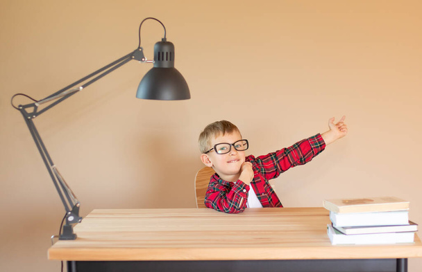 Um menino alegre com óculos senta-se em uma mesa e aponta o dedo para algo. De volta ao conceito de escola. Espaço de cópia - Foto, Imagem