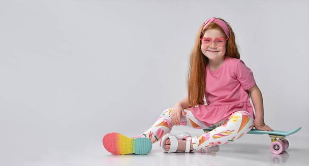 Smiling little red-haired girl child sitting on skateboard gesturing victory - Foto, Imagem