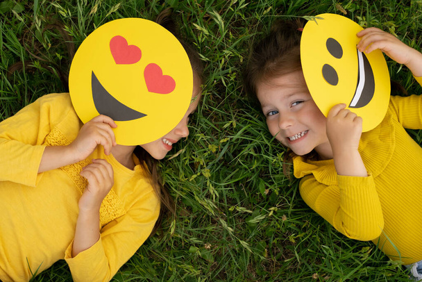 Little girls are lying on the lawn covering part of their faces with emoticons and laughing merrily. World smile Day. Anthropomorphic smile Face - Photo, Image