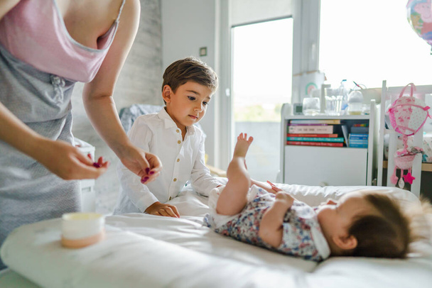 Pieni valkoihoinen poika nelivuotias auttaa äitiään vaihtamaan vaippoja pieneen pikkuveljeensä tai siskoonsa valoisassa makuuhuoneessa kotona perheen sitoutuminen ja hoito-konsepti. - Valokuva, kuva
