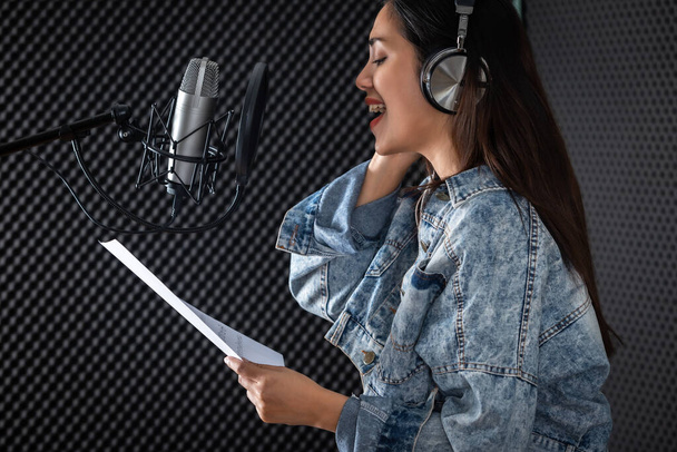 Счастливая веселая красивая улыбка портрета молодой азиатки-вокалистки, носящей наушники, записывающая песню перед микрофоном в профессиональной студии - Фото, изображение