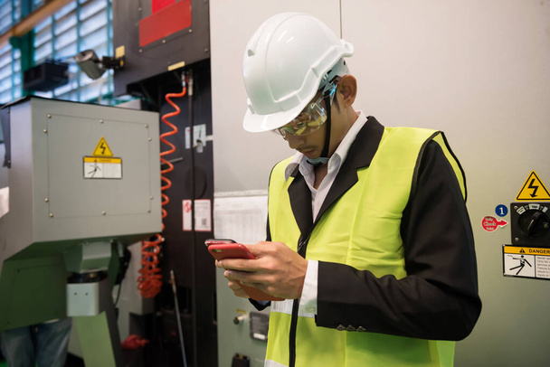 Busy factory Asian engineer manager checking manufacturing project plan from 2 smartphone devices near production machine with safety warning symbol. - Fotografie, Obrázek