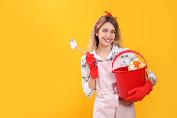 Young housewife with bucket of household items and brush on yellow background. Space for text - Fotoğraf, Görsel