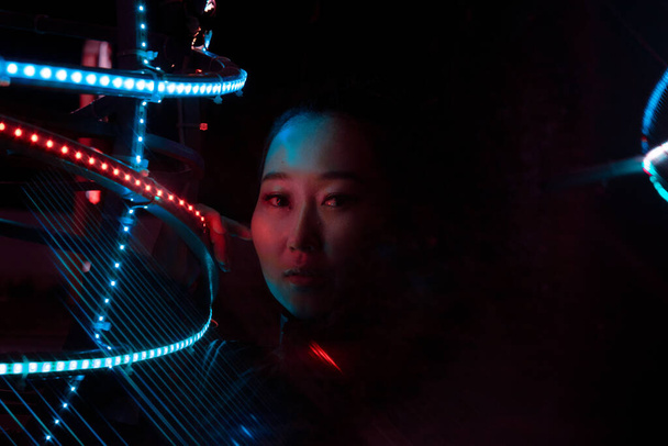 Portret dziewczyny w stylu cyberpunka, używającej refrakcji optycznej z użyciem pryzmatu. Nocne miasto, spojrzenie dziewczyny skierowane jest bezpośrednio na kamerę, jej ręka opiera się o źródło światła - Zdjęcie, obraz