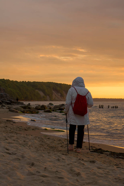 A man in a white jacket with a red backpack is engaged in Scandinavian walking in the evening on the seashore. - Photo, Image