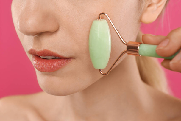 Young woman using natural jade face roller on pink background, closeup - Photo, Image