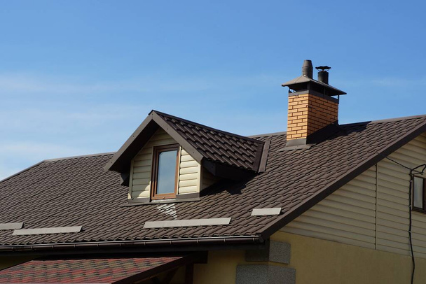 część prywatnego domu ze strychu z oknem pod brązowym dachem z ceglanym kominem na tle niebieskiego domu - Zdjęcie, obraz