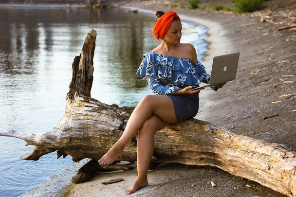 Eine junge schöne muslimische malaiische Geschäftsfrau mit orangefarbenem Hidschab-Turban auf dem Kopf, die an einem Sommertag am Meer am Laptop sitzt. Fernarbeit, Freizeit, Reisekonzept. Videokonferenzen online. - Foto, Bild