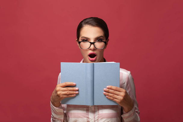 Portrait of smart surprised woman in glasses holding book with empty cover posing isolated on red wall background. Mock up copy space. People sincere emotions lifestyle concept - Photo, Image