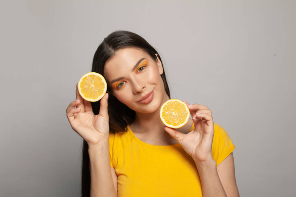 Linda mujer sonriente en camiseta amarilla sosteniendo frutas de limón. Bonita modelo femenina con piel clara y cabello castaño liso sobre fondo blanco - Foto, imagen