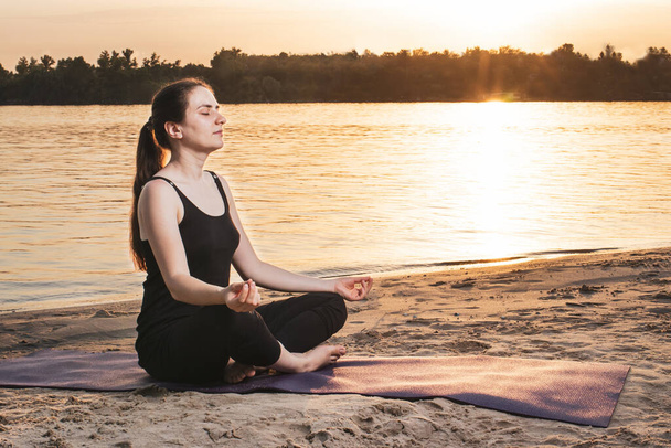 A woman meditates and does yoga on the river bank at sunset. Yoga in nature and unity with nature place for text. - Zdjęcie, obraz