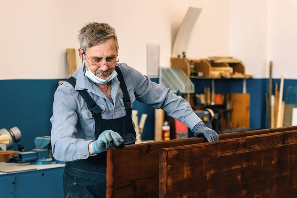 Starší muž maluje dřevěné desky s tmavým lakem v truhlářské dílně. Důchodce v lékařské masce  - Fotografie, Obrázek