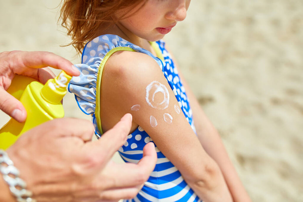 Vader brengt zonnebrandcrème aan op dochter op het strand, vader schildert zonnebrandcrème op schouder in de vorm van de zon, beschermt de huid van de zonnecrème. Zomervakantie. - Foto, afbeelding