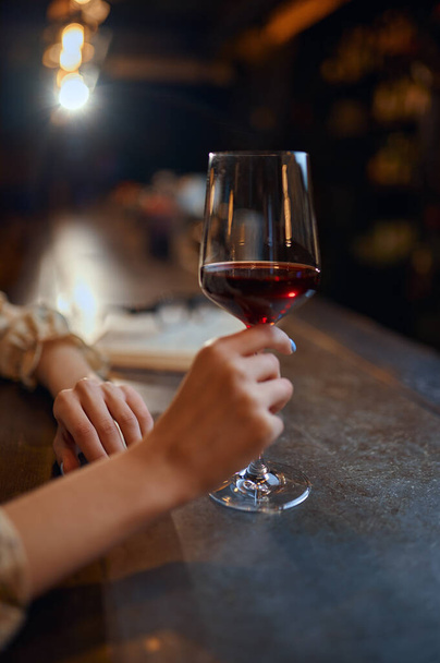 Γυναίκα με ένα ποτήρι κόκκινο κρασί κάθεται στον πάγκο στο μπαρ. Ένα θηλυκό πρόσωπο σε παμπ, ανθρώπινα συναισθήματα, δραστηριότητες αναψυχής, νυχτερινή ζωή - Φωτογραφία, εικόνα