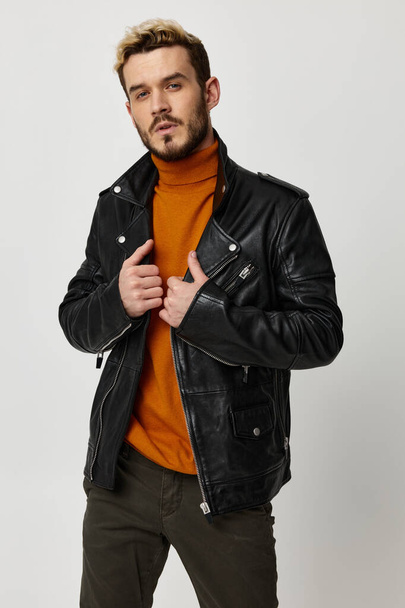 a man in an orange sweater straightens a leather jacket on a light background and fashionable trousers - Фото, изображение