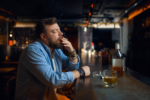 バーのカウンターでストレスをかけられた髭の男が酒を飲む。パブで休む一人の男性、人間の感情やレジャー活動、うつ病、ストレス軽減 - 写真・画像
