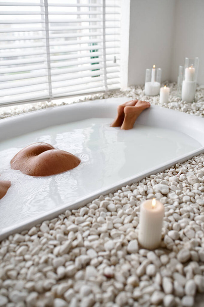Young sexy woman, relaxation in bath with milk, view on buttocks. Female person in bathtub, beauty and health care in spa, wellness treathment in bathroom, pebbles and candles on background - Photo, Image