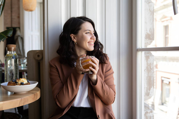 femme souriante assise dans un café à boire du thé en regardant dans l'espace de copie de la fenêtre - Photo, image