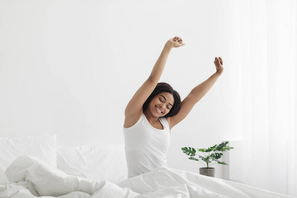 Happy black woman full of vitality and energy, waking up and stretching hands, sitting on bed and smiling, copy space - Foto, imagen