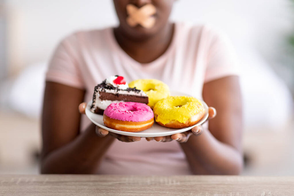 Крупный план кривой черной леди с тарелкой сладостей, с клейкой повязкой на рту, придерживаясь диеты потери веса - Фото, изображение