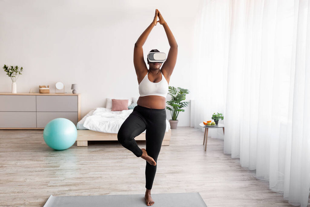 Overweight senhora negra vestindo fone de ouvido VR, praticando ioga em realidade aumentada em casa - Foto, Imagem