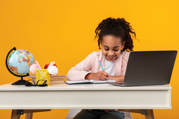 Πορτρέτο του χαμογελαστού μαύρου κοριτσιού που κάθεται στο γραφείο και γράφει - Φωτογραφία, εικόνα