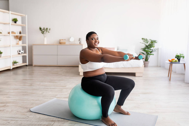 Grande taille femme noire travaillant avec des haltères sur balle de fitness à la maison, espace vide - Photo, image