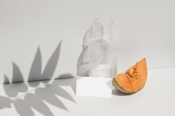 Summer food still life composition. Cantaloupe melon fruit on table. White wall with palm leaf shadow overlay. Blank paper card, invitation mockup. Glass carafe of water, cocktail in sunlight. - Photo, image
