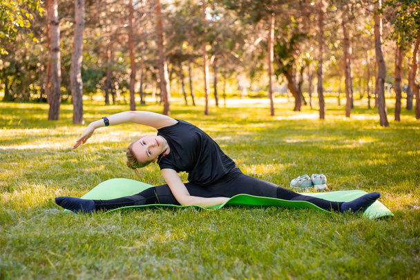 Outdoor Pilates training. A young woman does exercises to stretch the muscles of her legs while sitting on a yoga mat in the park. Exercise for the development of twine. - Foto, Imagen