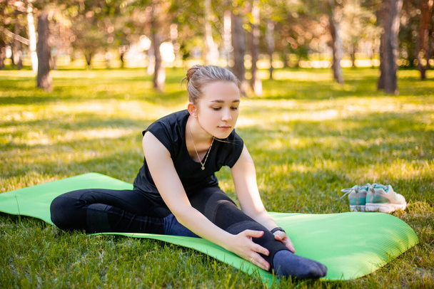 Outdoor Pilates training. A young woman does exercises to stretch the muscles of her legs while sitting on a yoga mat in the park. Exercise for the development of twine. - Foto, Imagen