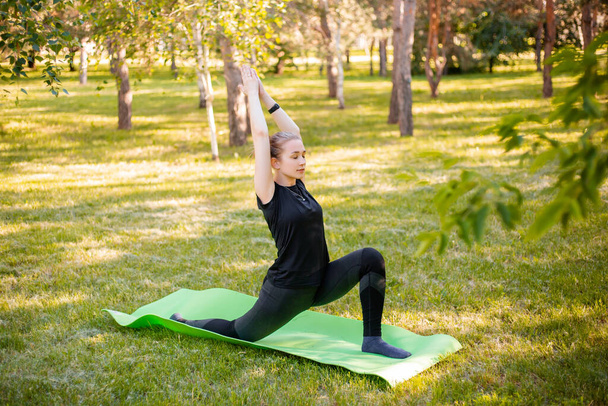 Outdoor Pilates training. A young woman does exercises to stretch the muscles of her legs while sitting on a yoga mat in the park. Exercise for the development of twine. - Φωτογραφία, εικόνα