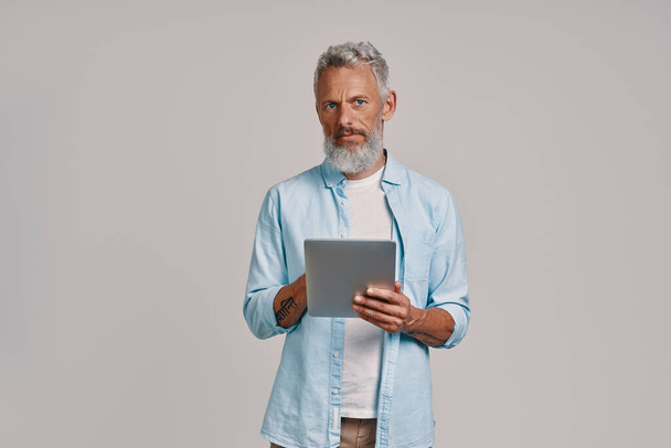 Uomo anziano fiducioso utilizzando tablet digitale mentre in piedi su sfondo grigio - Foto, immagini