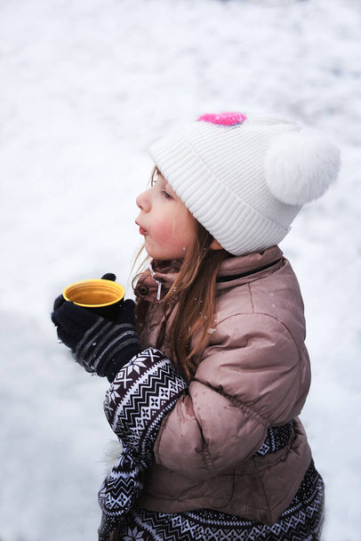 Child drinks hot tea in park in winter. Cute little Caucasian girl in warm knitted hat and jacket holds cup of tea in her hands and warms up in cold weather. Mittens on kids hands and warming drink. - Photo, Image