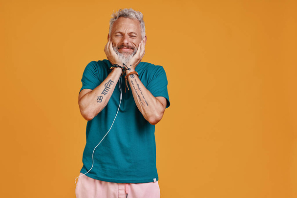 Happy senior man listening music and smiling while standing against orange background - Foto, afbeelding