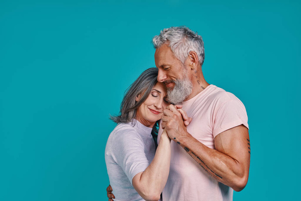 Romantic senior couple dancing and smiling while standing together against blue background - Zdjęcie, obraz