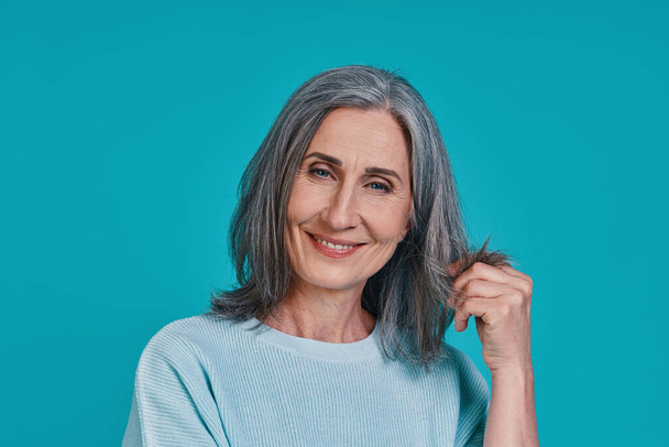 Headshot of mature beautiful woman looking at camera and smiling while standing against blue background - Foto, Imagem