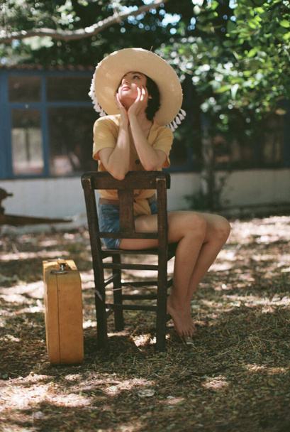 Country woman sits on a wooden chair with a straw hat yellow shirt and jean shorts. Shot with an analog camera and 35mm film.. Film grain intentionally not removed - Foto, immagini