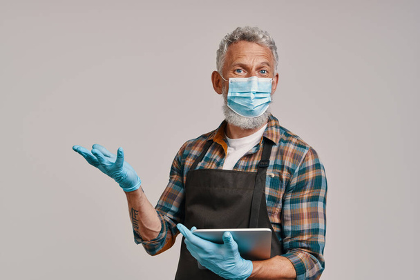Senior man in apron and protective face mask holding digital tablet and gesturing - Foto, Bild