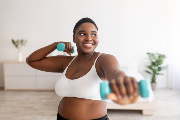 Οικιακά αθλήματα για την απώλεια βάρους. Ευτυχισμένο συν μέγεθος μαύρη γυναίκα κάνει ασκήσεις γυμναστικής με αλτήρες σε εσωτερικούς χώρους - Φωτογραφία, εικόνα