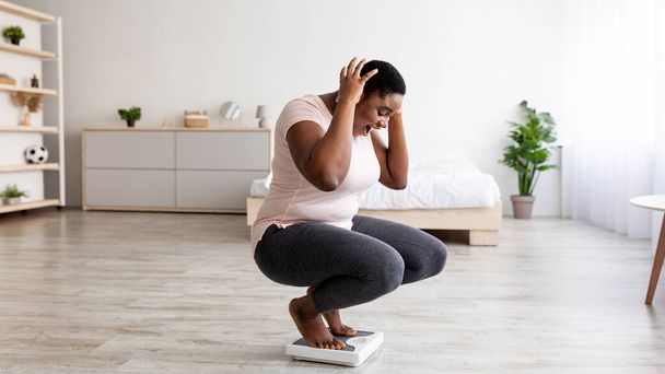 Além de tamanho mulher negra não pode acreditar em seu resultado de perda de peso, gritando OMG, sentado em escalas em casa, sentindo-se animado - Foto, Imagem